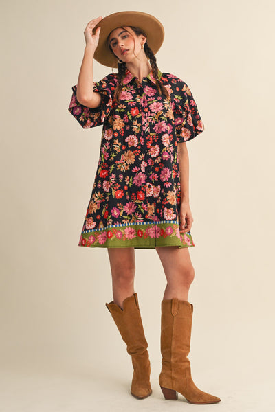A woman modeling a short-sleeve black dress with a vibrant multicolor floral print and a green patterned hem, styled with a tan wide-brim hat and brown suede knee-high boots against a solid beige background.