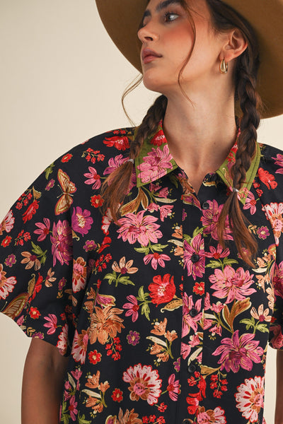 A close-up of a woman wearing a short-sleeve button-up shirt with a vibrant pink, red, and orange floral print on a black background, paired with a wide-brimmed tan hat.