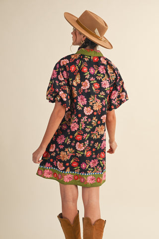 A woman shown from the back wearing a short-sleeve black floral dress with a green collar and hem, a tan hat, and brown boots, against a plain beige studio background.