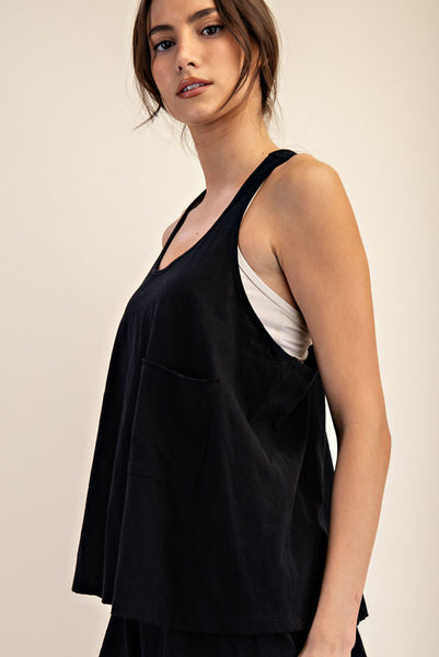 A woman wearing a black sleeveless tank top with a front pocket, layered over a white camisole, photographed against a plain light beige studio background.