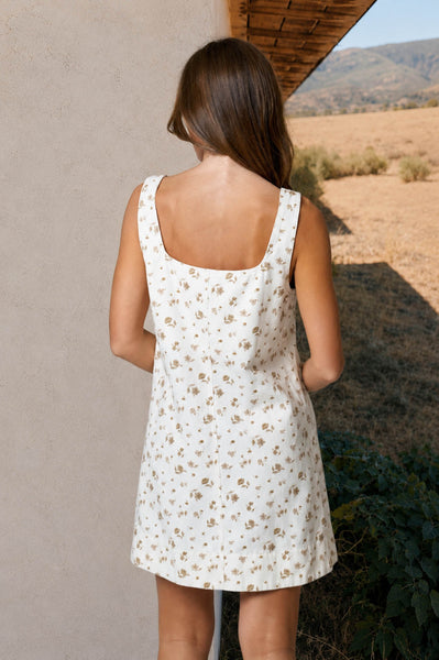 Woman wearing a white floral dress standing against a beige wall with a desert landscape in the background.