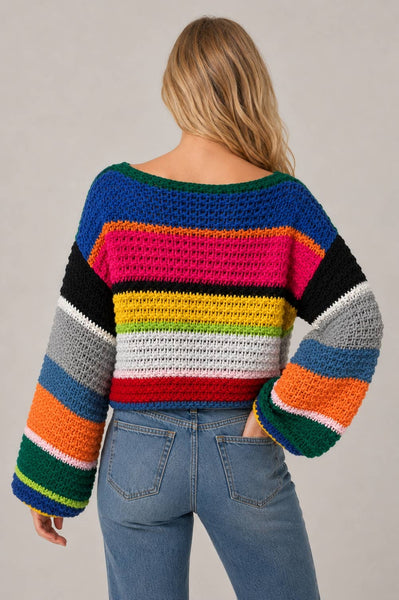 A woman shown from the back wearing a colorful striped crocheted sweater and blue jeans against a plain light gray studio background.