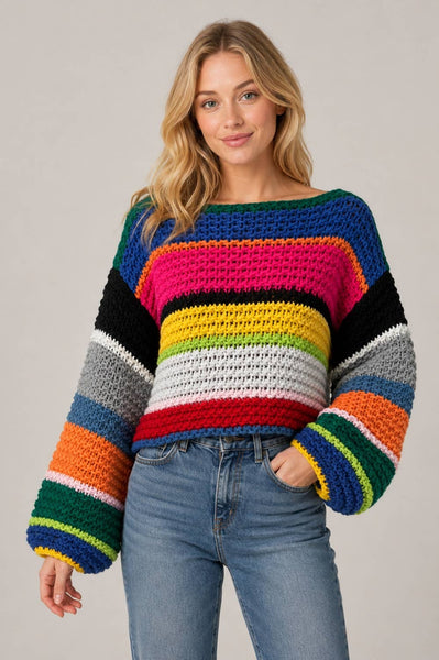 A woman wearing a chunky crocheted sweater with bold horizontal rainbow stripes in blue, pink, black, yellow, green, orange, white, and gray, paired with light-wash blue jeans, against a solid light gray background.