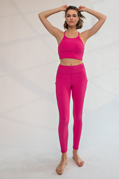A woman modeling a bright pink activewear set consisting of a sports bra and high-waisted leggings, standing against a light gray studio background.