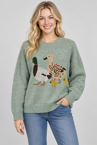 A woman wearing a sage green knit sweater with a large duck and duckling graphic across the chest, paired with blue jeans, against a solid light gray background.