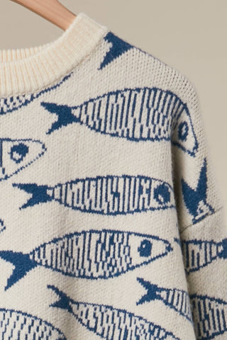 Close-up of a cream knit sweater with a repeating blue fish pattern, hanging on a wooden hanger against a light beige background.