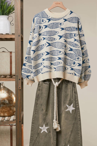 A cream-colored knit sweater with a blue fish pattern, styled on a wooden hanger with an olive green skirt featuring white star patches and a rope belt, set against a rustic wooden shelf background.