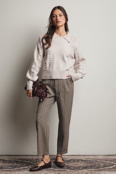 Woman wearing a light-colored sweater and plaid pants, holding a brown bag against a plain background