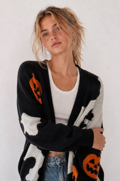 A woman wearing a black knit cardigan with orange jack-o'-lantern motifs and white ghost-like patches, layered over a white ribbed tank top and blue jeans, against a plain light background.