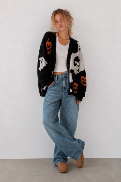 A woman modeling a black knit cardigan with white ghost and orange pumpkin motifs, worn over a white crop top and blue wide-leg jeans.