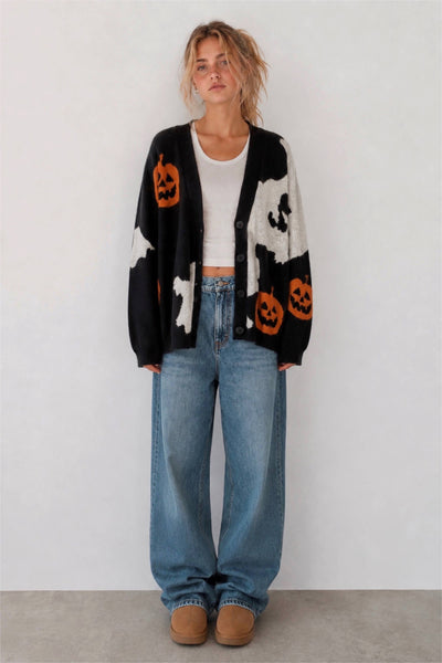 A woman stands against a plain light gray wall wearing a black cardigan with orange jack-o'-lantern motifs and white ghost patterns, over a white cropped top and loose blue jeans.