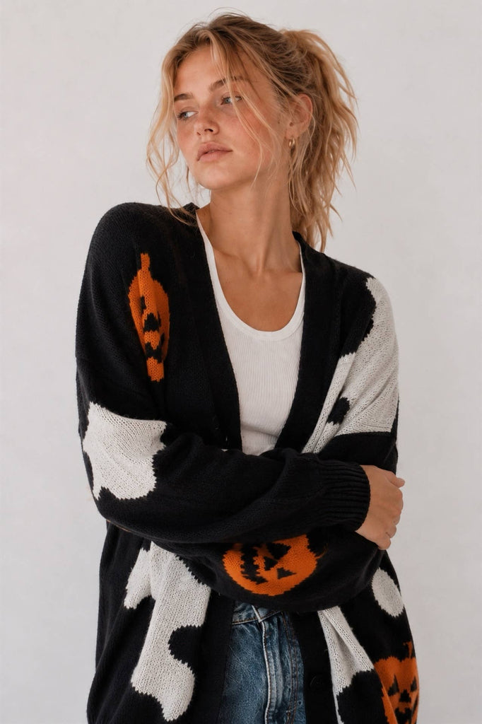 A woman wearing a black and white cow-print cardigan with orange pumpkin motifs, layered over a white t-shirt and blue jeans, photographed against a plain light gray background.