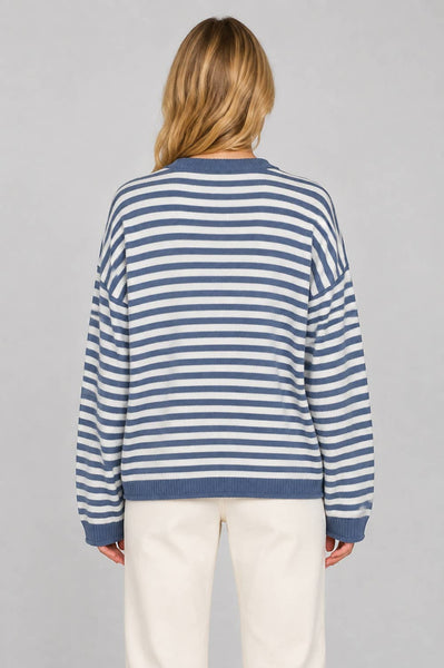 A woman viewed from behind wearing a blue and white horizontally striped knit sweater and cream-colored pants, set against a solid light gray studio background.