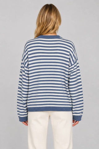 A woman viewed from behind wearing a blue and white horizontally striped knit sweater and cream-colored pants, set against a solid light gray studio background.