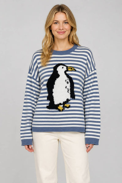 A woman wearing a blue and white striped knit sweater with a large penguin motif on the front, paired with cream pants, against a solid gray studio background.