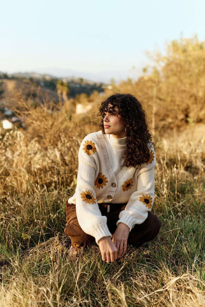 Woman sitting in a field wearing a white sweater with sunflower patterns