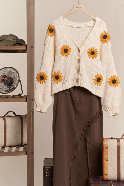 White cardigan with sunflower patterns on a hanger against a neutral background.