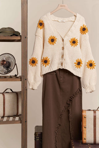 White cardigan with sunflower patterns on a hanger against a neutral background.