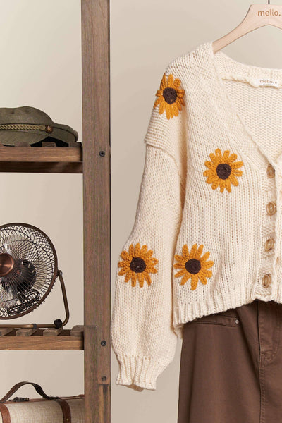 Beige cardigan with sunflower patterns hanging on a wooden rack.