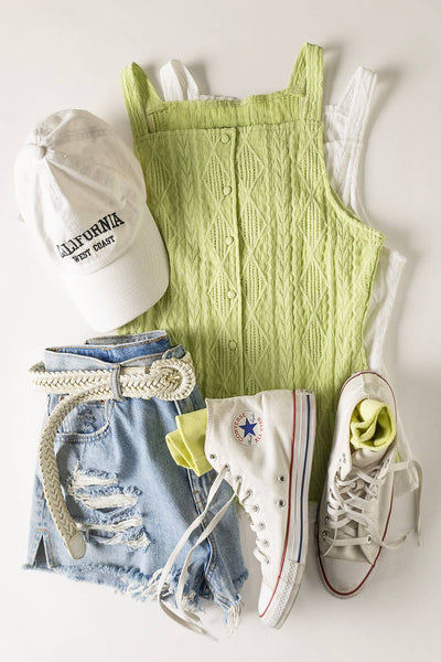 Green knitted romper with white sneakers, denim shorts, and a white cap on a light background