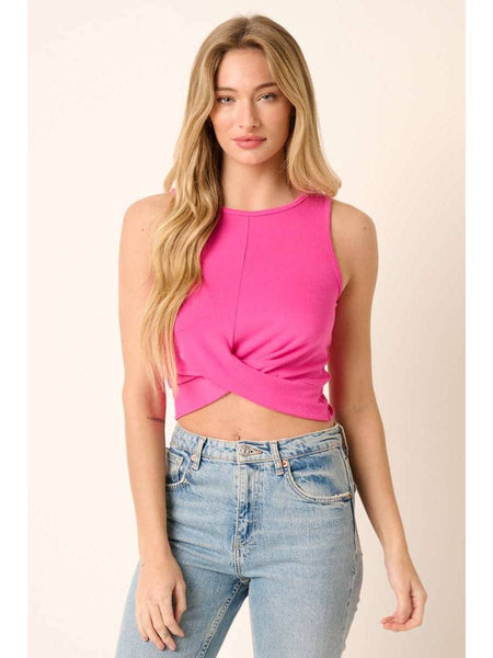 A woman modeling a sleeveless bright pink crop top with a twisted front detail, paired with light-wash blue jeans, against a solid light beige background.