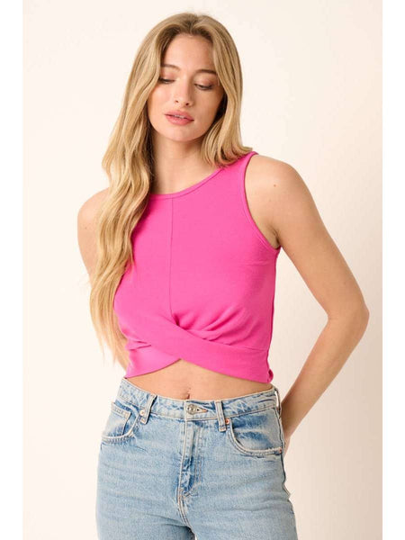 A woman wearing a bright pink sleeveless crop top with a twisted front detail and light-wash blue jeans, standing against a plain light beige background.