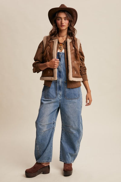 Woman wearing a brown jacket, beige vest, blue jeans, and brown hat on a beige background