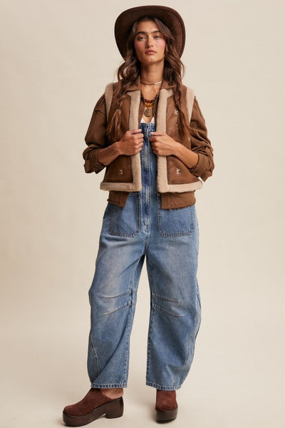 Woman wearing a brown jacket, denim overalls, and a hat on a beige background