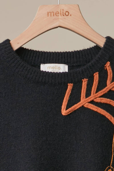 A close-up of a black knit sweater hanging on a light wood hanger branded 'mello.' The sweater has a ribbed crew neck and a prominent orange embroidered abstract leaf or branch motif on the right side.
