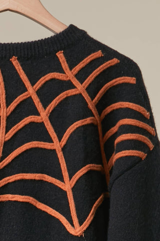 Close-up of a black knit sweater with a large orange spiderweb pattern across the chest and shoulders, hanging on a wooden hanger against a plain light beige background.