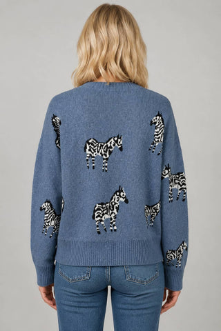A woman viewed from behind wearing a heathered blue knit sweater with black and white zebra motifs across the back, paired with blue jeans, against a neutral gray studio background.