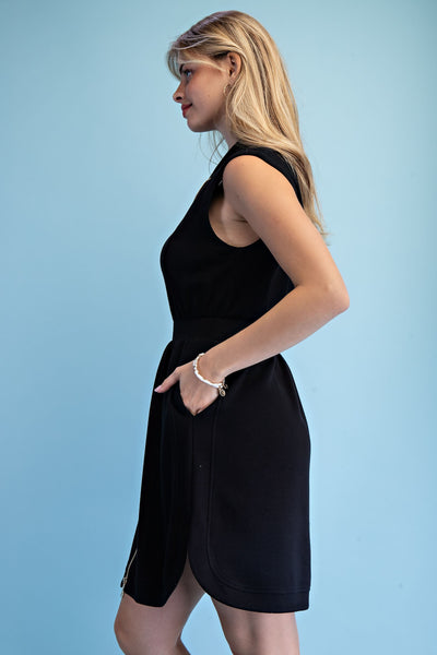 A woman in a side profile wearing a sleeveless black dress with a wrap-style hem, standing against a solid light blue background.